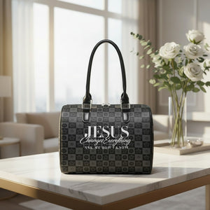 A black and gray patterned handbag with the text 'JESUS Change Everything' printed on the front, displayed on a marble table in a bright, modern living room with white roses in the background.